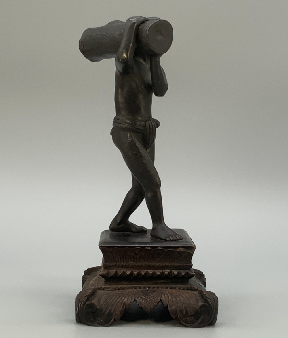 A dark patinated bronze figurine of a man carrying a log on his shoulder, mounted on an intricately carved wooden stand. View VIII