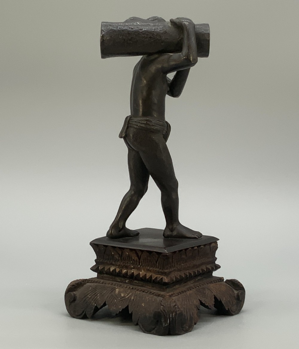 A dark patinated bronze figurine of a man carrying a log on his shoulder, mounted on an intricately carved wooden stand. View IX