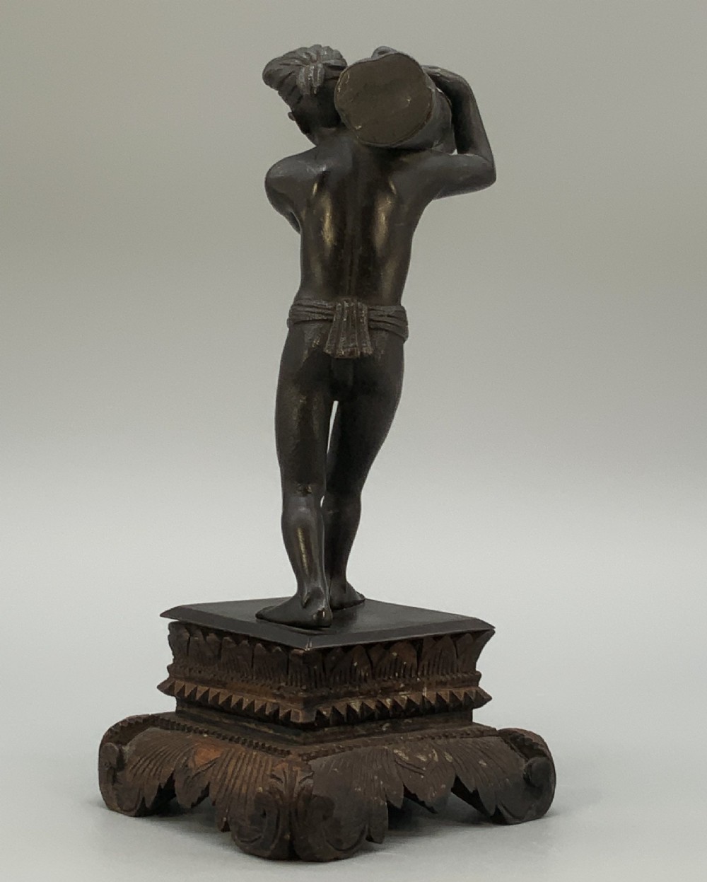 A dark patinated bronze figurine of a man carrying a log on his shoulder, mounted on an intricately carved wooden stand. View X