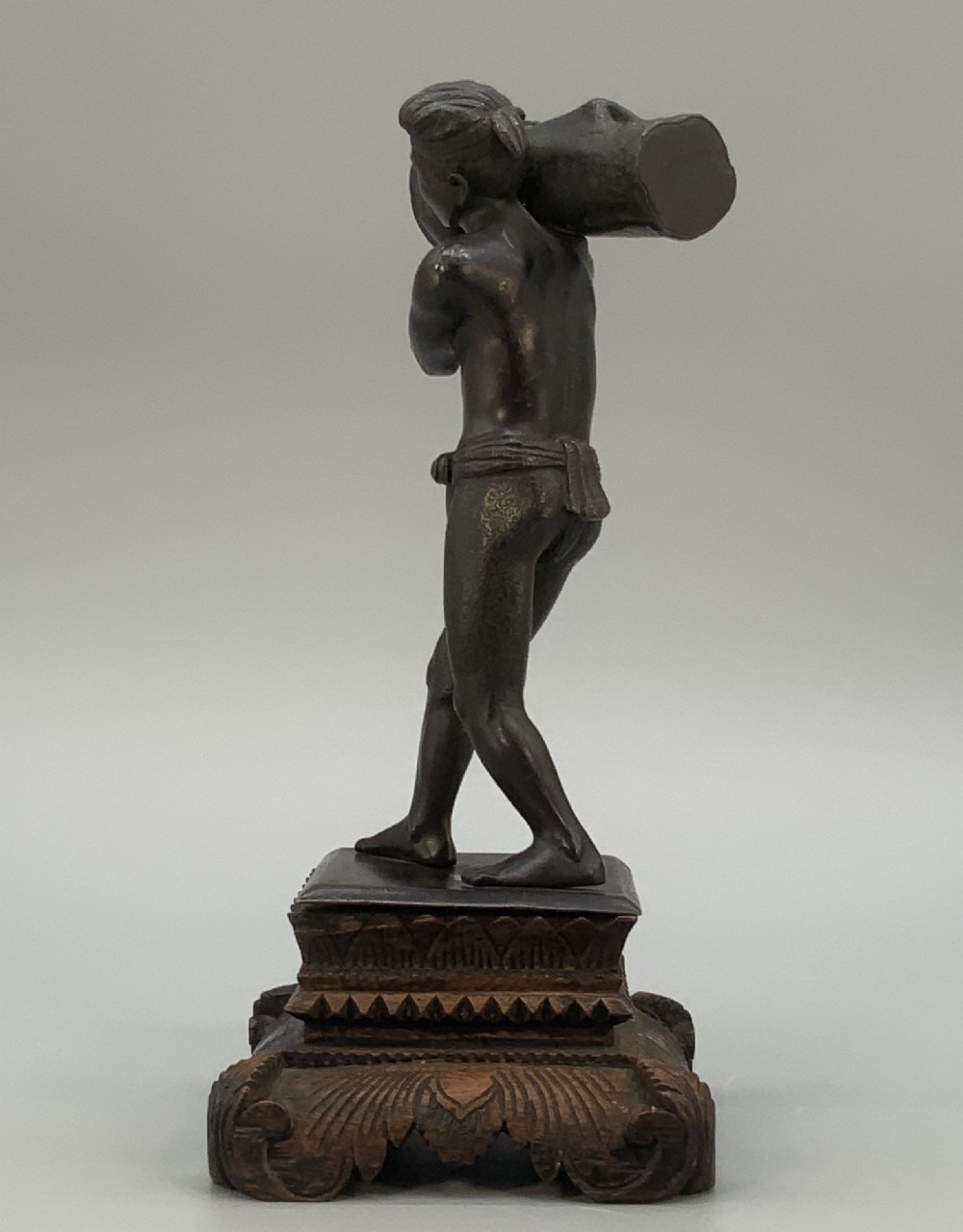 A dark patinated bronze figurine of a man carrying a log on his shoulder, mounted on an intricately carved wooden stand. View XI
