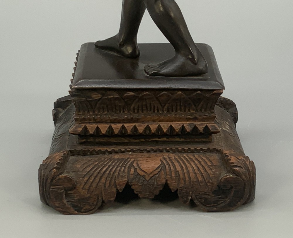 A dark patinated bronze figurine of a man carrying a log on his shoulder, mounted on an intricately carved wooden stand. View XII