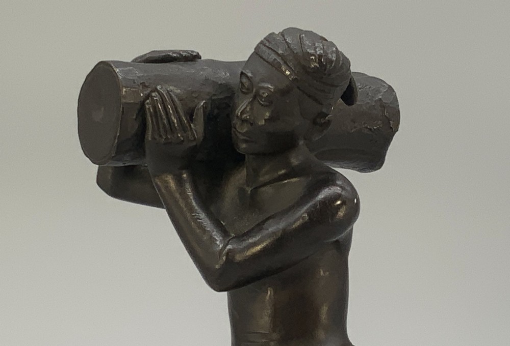 A dark patinated bronze figurine of a man carrying a log on his shoulder, mounted on an intricately carved wooden stand. View XIII