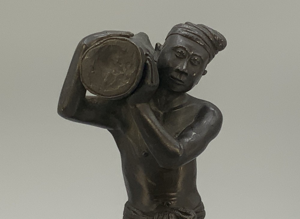 A dark patinated bronze figurine of a man carrying a log on his shoulder, mounted on an intricately carved wooden stand. View XIV