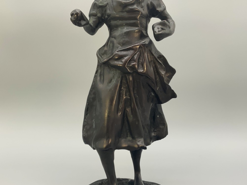 An antique dark bronze sculpture of a young maiden wearing a draped apron dress, standing barefoot on a rounded integral base. View V