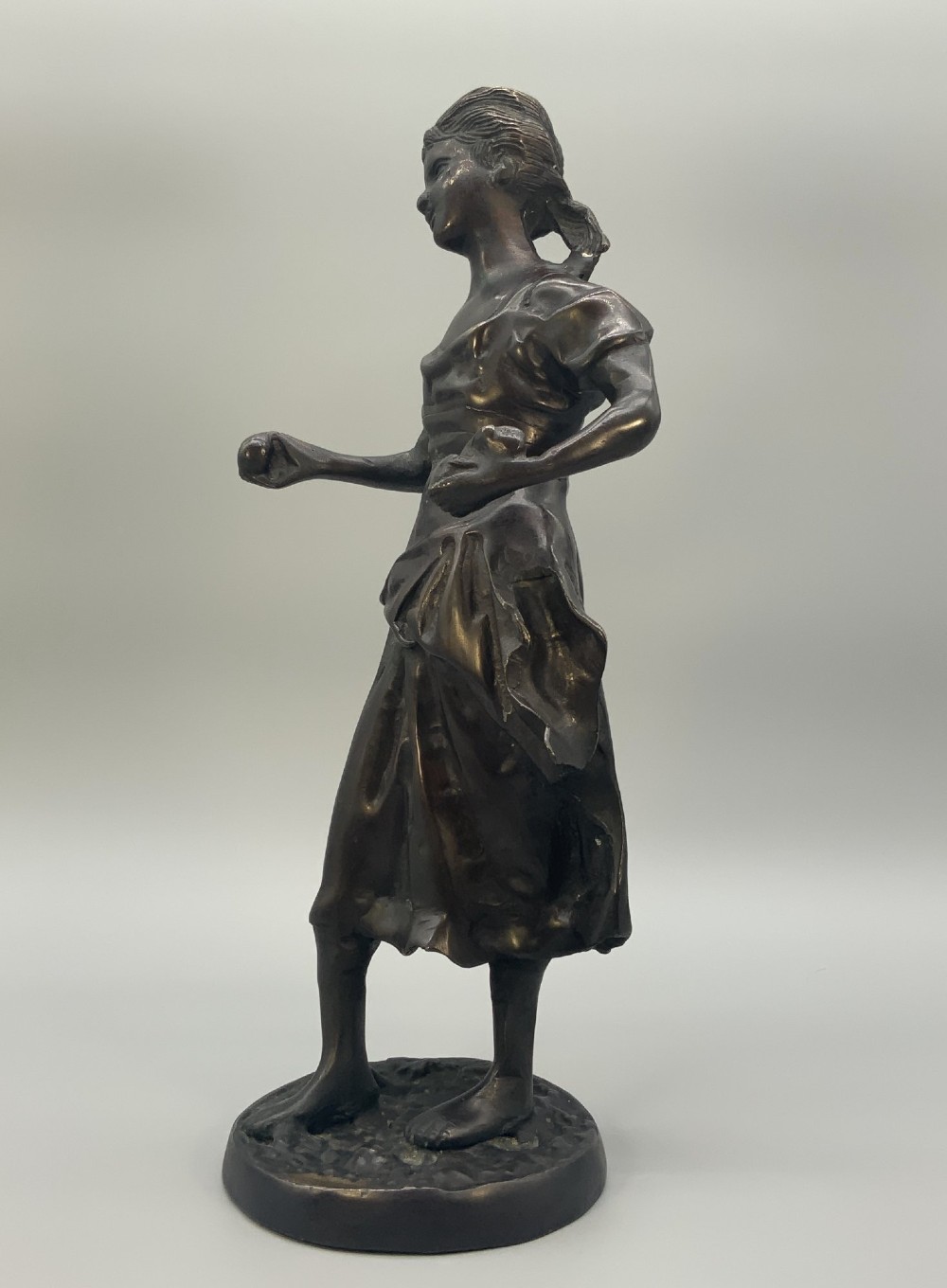 An antique dark bronze sculpture of a young maiden wearing a draped apron dress, standing barefoot on a rounded integral base. View VII