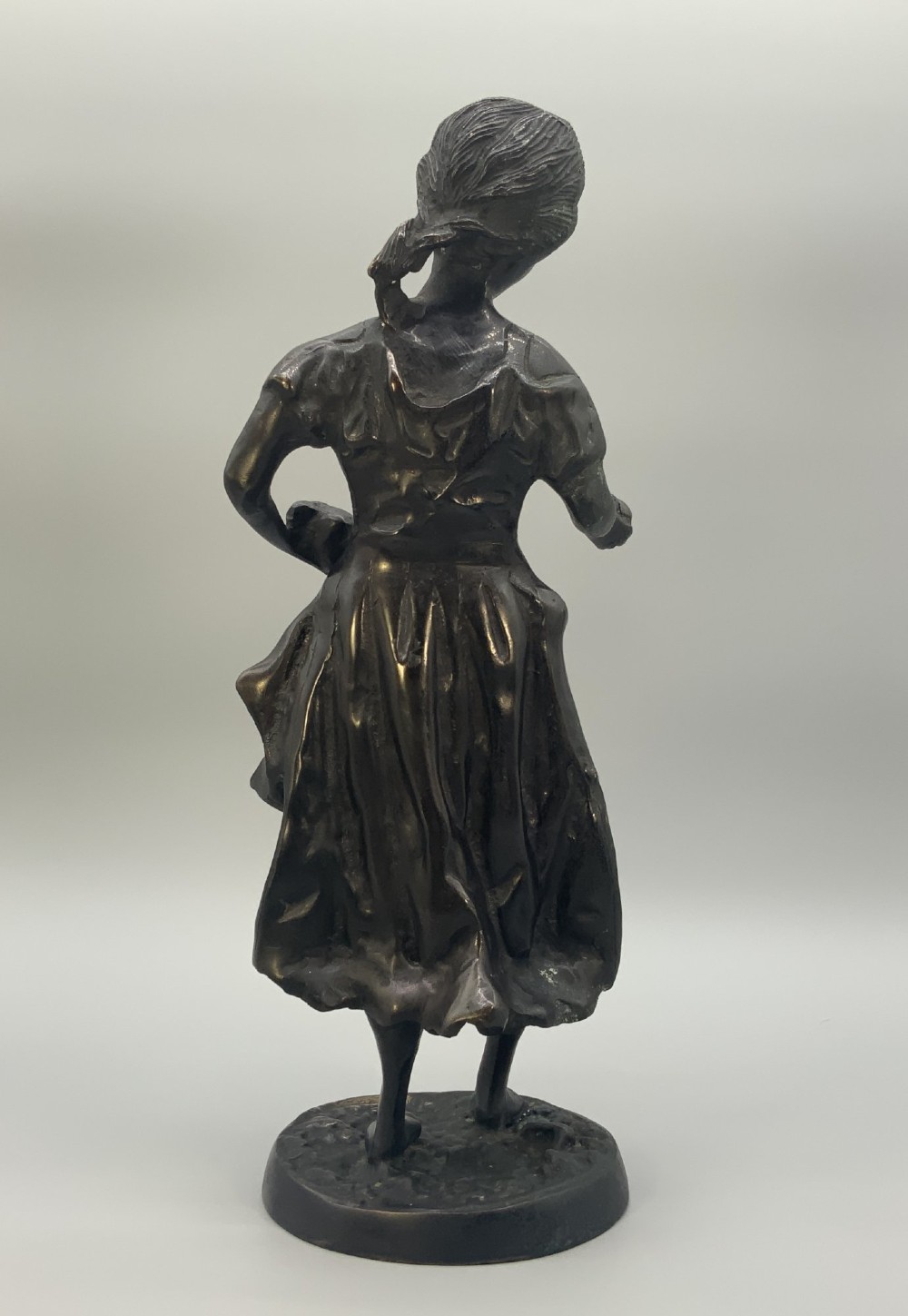 An antique dark bronze sculpture of a young maiden wearing a draped apron dress, standing barefoot on a rounded integral base. View VIII
