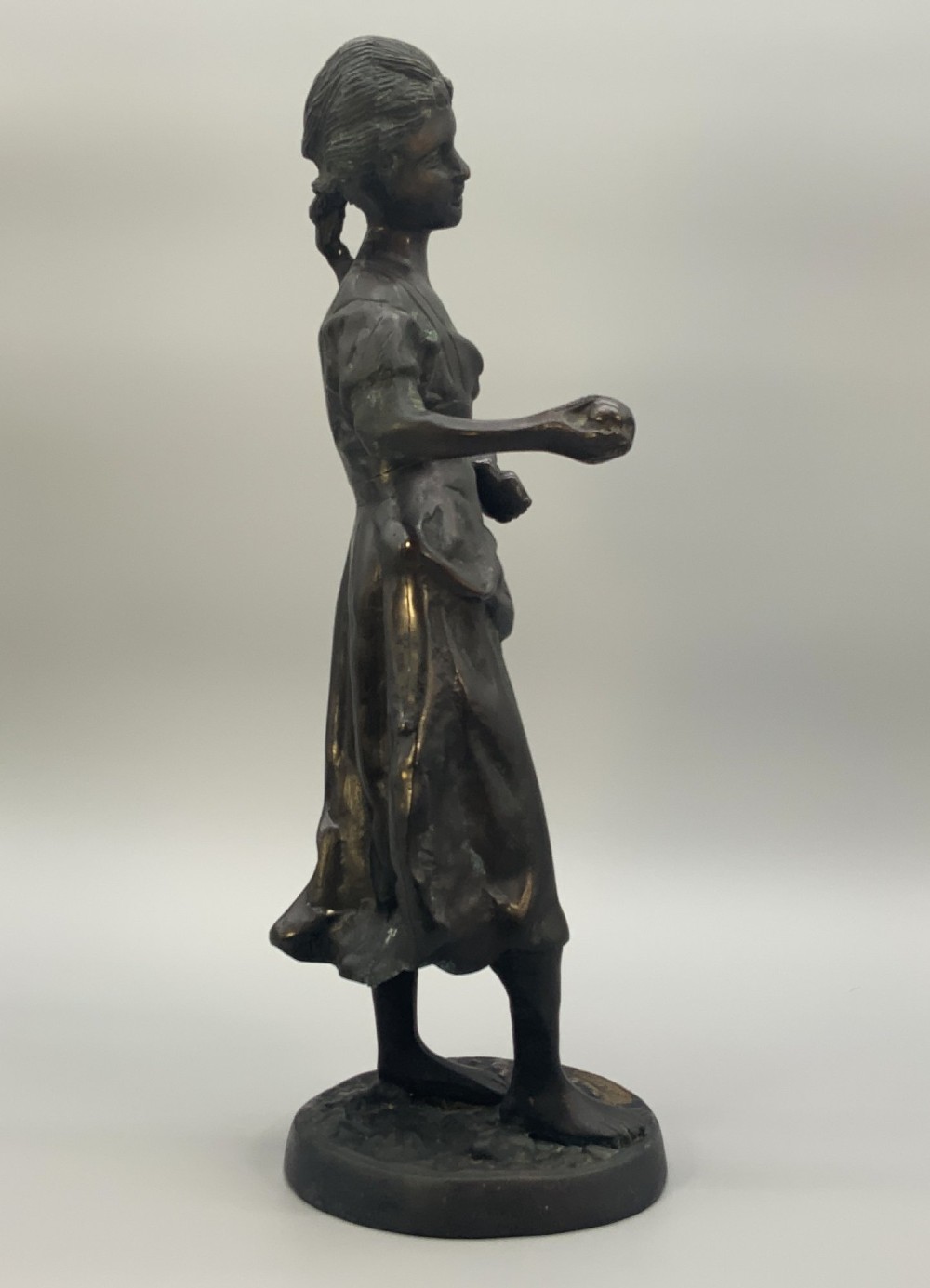 An antique dark bronze sculpture of a young maiden wearing a draped apron dress, standing barefoot on a rounded integral base. View IX
