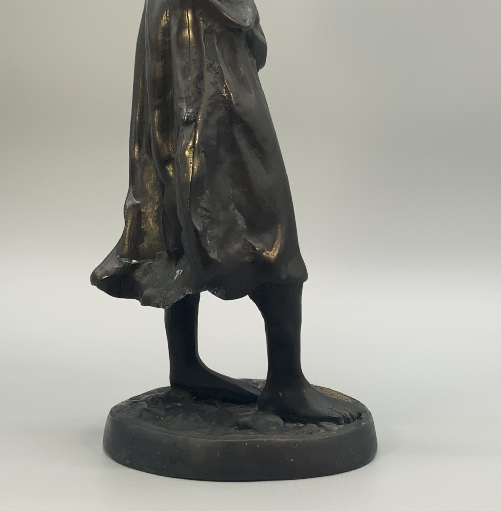 An antique dark bronze sculpture of a young maiden wearing a draped apron dress, standing barefoot on a rounded integral base. View XI