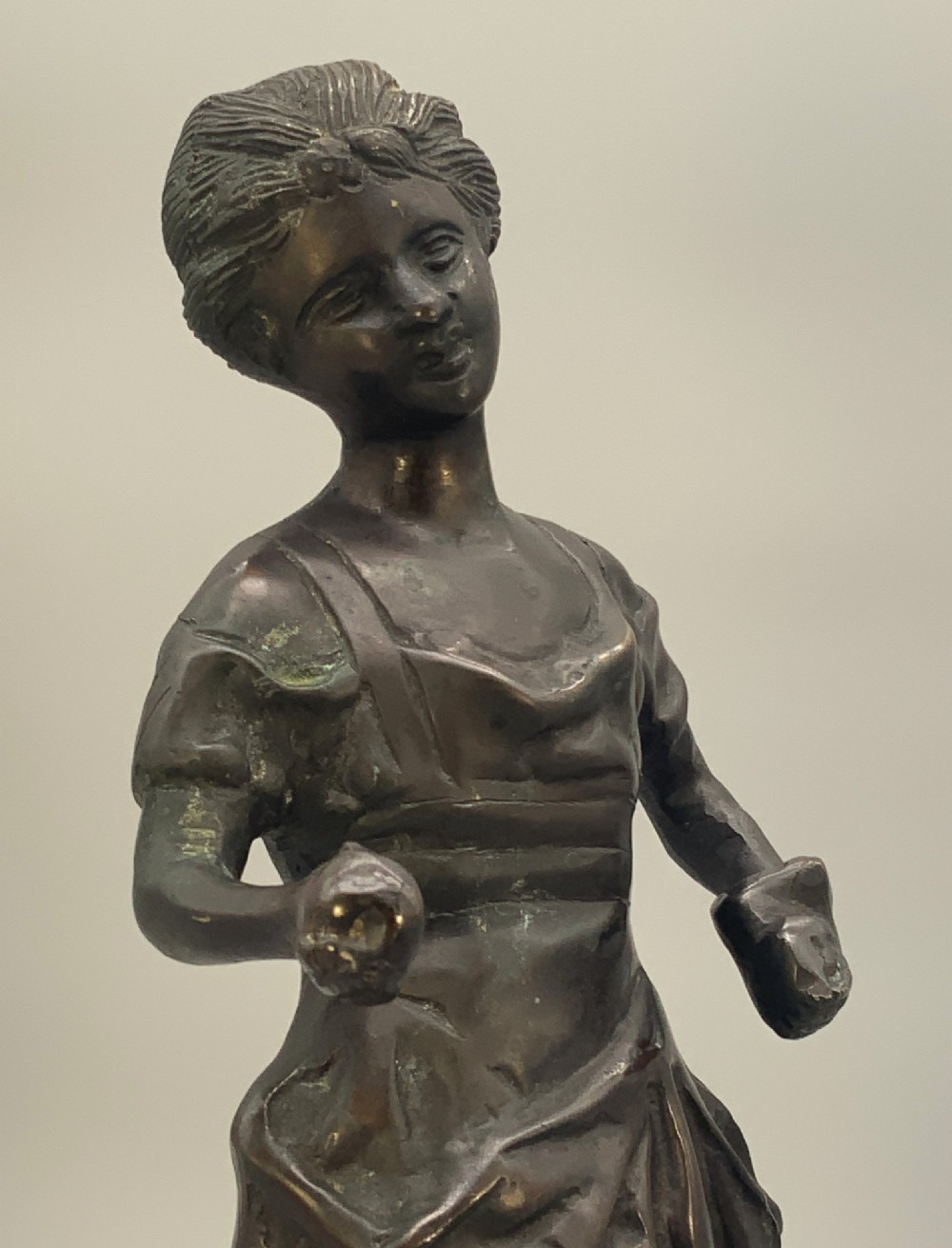An antique dark bronze sculpture of a young maiden wearing a draped apron dress, standing barefoot on a rounded integral base. View XII