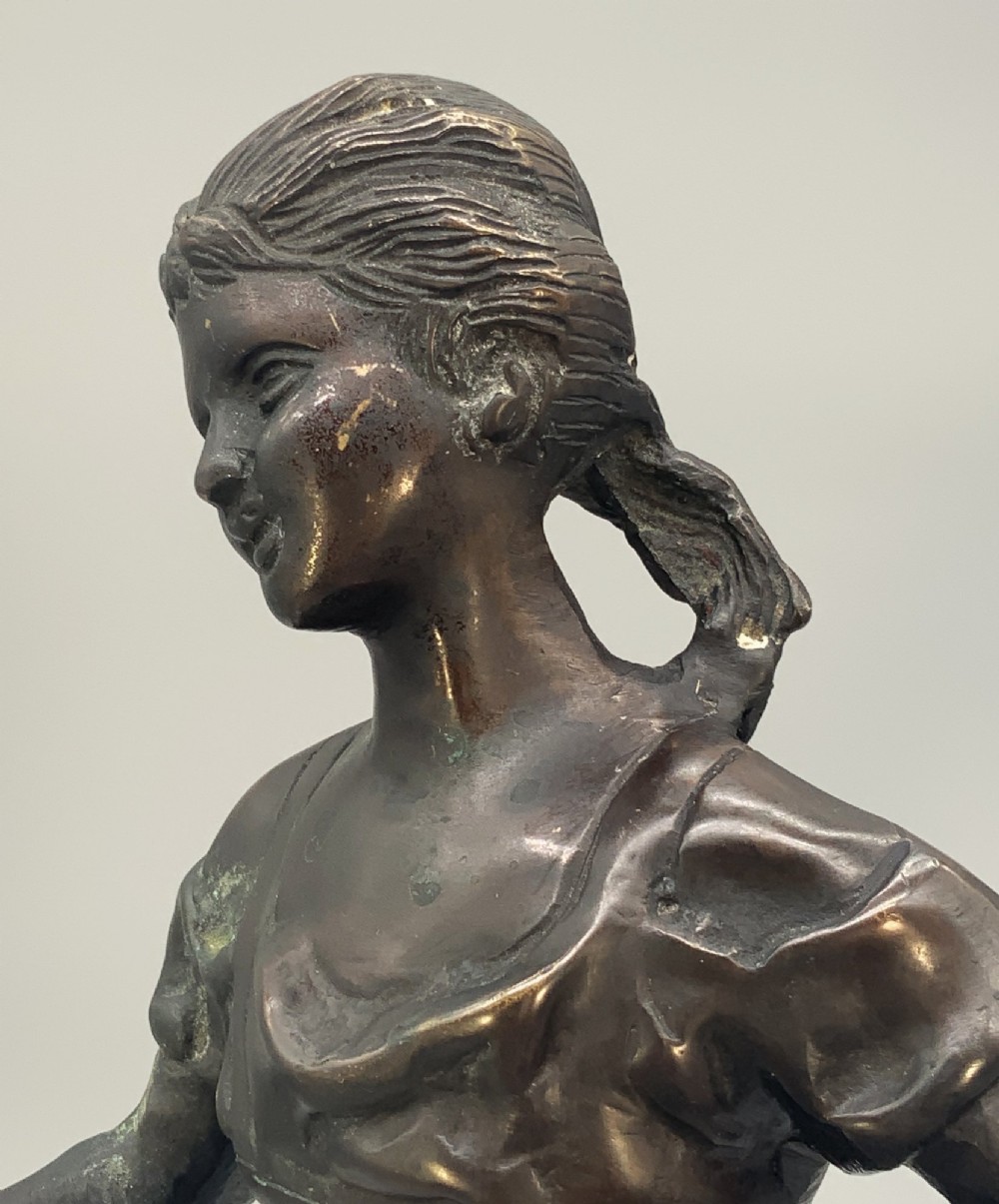 An antique dark bronze sculpture of a young maiden wearing a draped apron dress, standing barefoot on a rounded integral base. View XIV