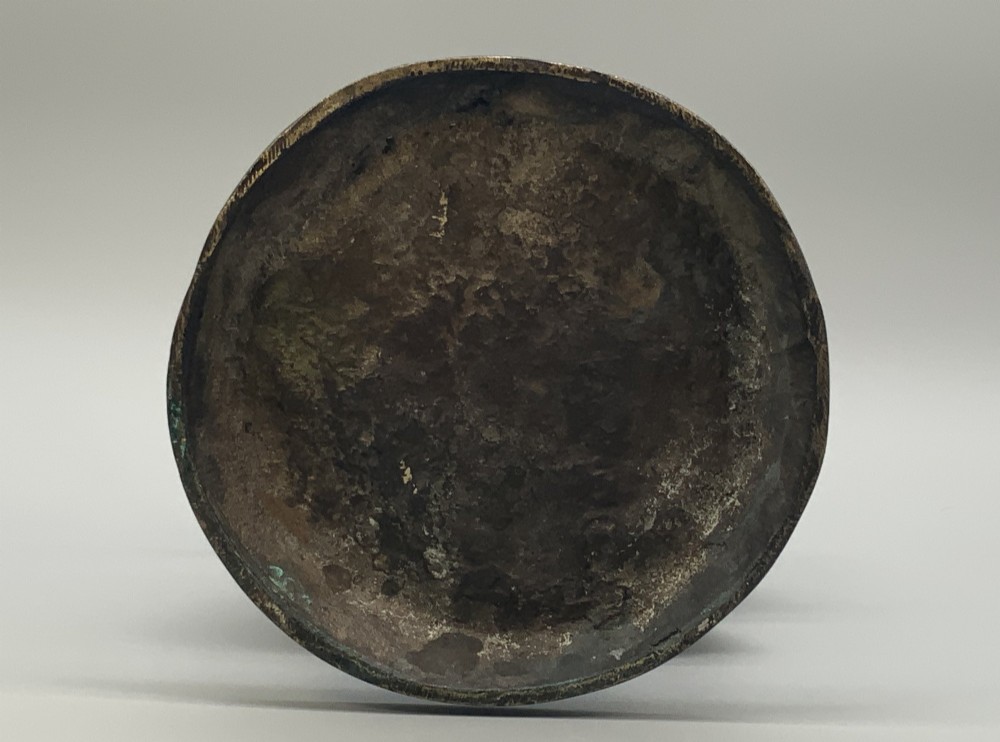 An antique dark bronze sculpture of a young maiden wearing a draped apron dress, standing barefoot on a rounded integral base. View XV