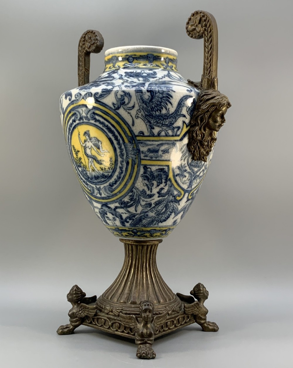 A Renaissance Revival faience vase featuring blue and yellow glazed classical medallion artwork mounted on a cast bronze footed base with mask handles. View VI