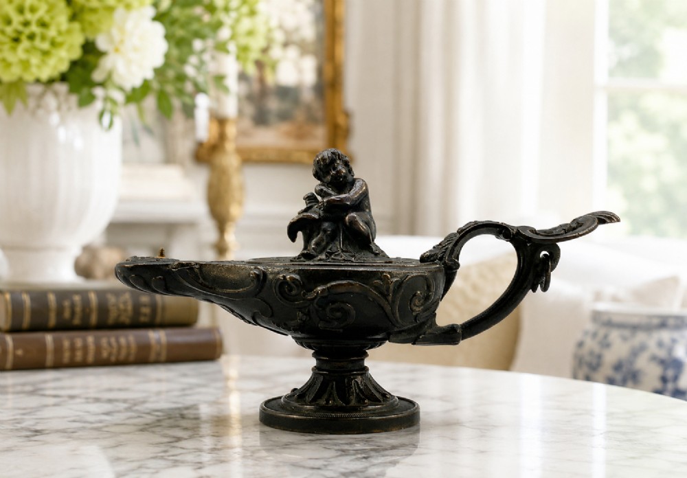 A patinated dark bronze classical oil lamp featuring a seated putto figure on top and a scroll handle resting on a pedestal base. View II