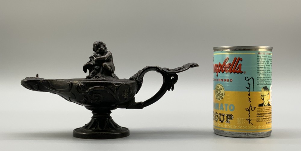 A patinated dark bronze classical oil lamp featuring a seated putto figure on top and a scroll handle resting on a pedestal base. View III