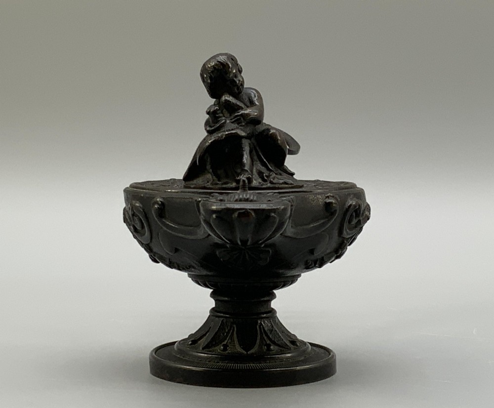 A patinated dark bronze classical oil lamp featuring a seated putto figure on top and a scroll handle resting on a pedestal base. View IV