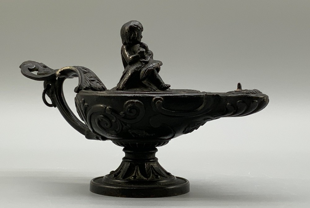 A patinated dark bronze classical oil lamp featuring a seated putto figure on top and a scroll handle resting on a pedestal base. View V