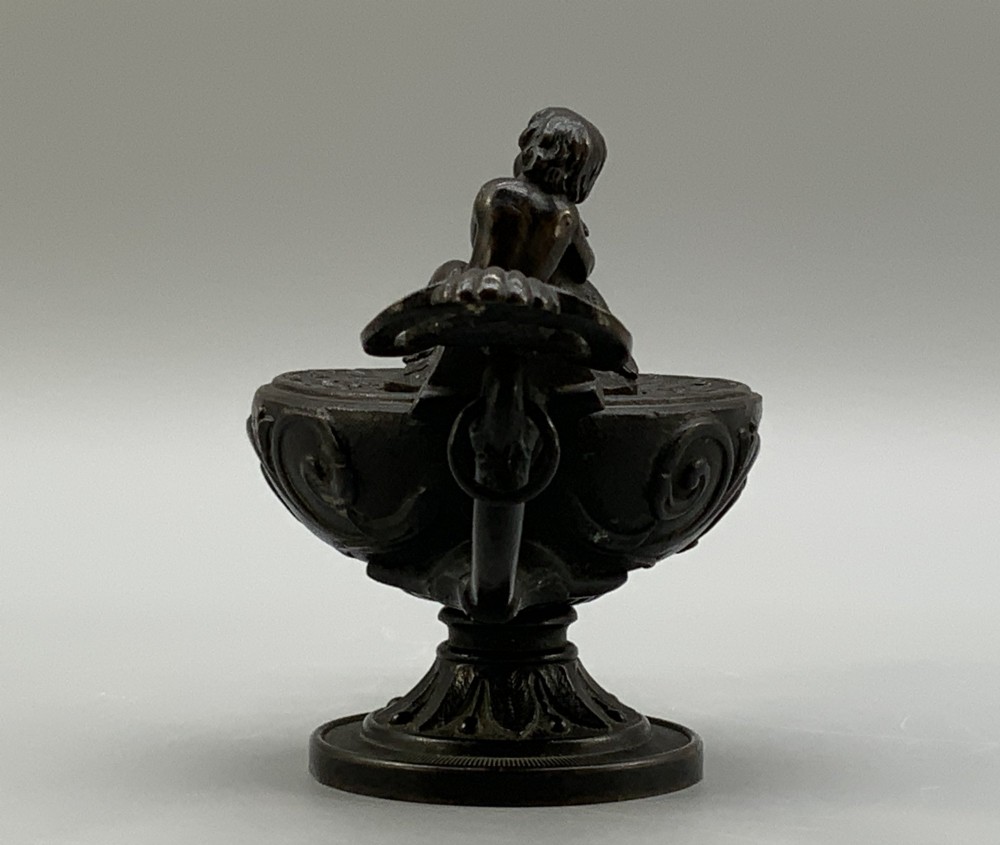 A patinated dark bronze classical oil lamp featuring a seated putto figure on top and a scroll handle resting on a pedestal base. View VI