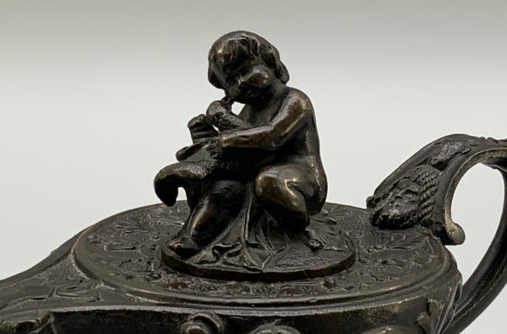 A patinated dark bronze classical oil lamp featuring a seated putto figure on top and a scroll handle resting on a pedestal base. View VII