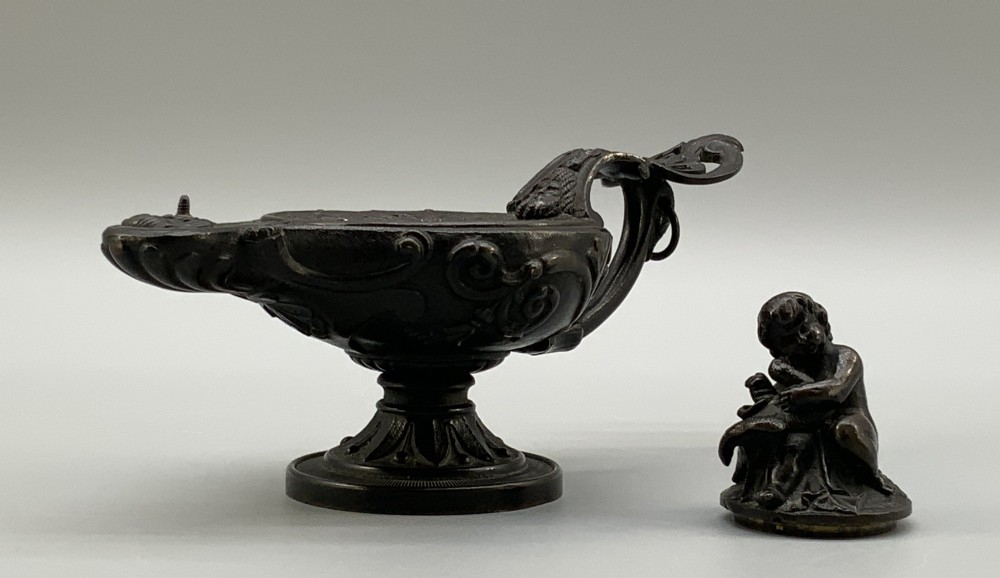 A patinated dark bronze classical oil lamp featuring a seated putto figure on top and a scroll handle resting on a pedestal base. View VIII