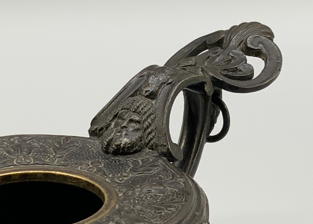 A patinated dark bronze classical oil lamp featuring a seated putto figure on top and a scroll handle resting on a pedestal base. View IX