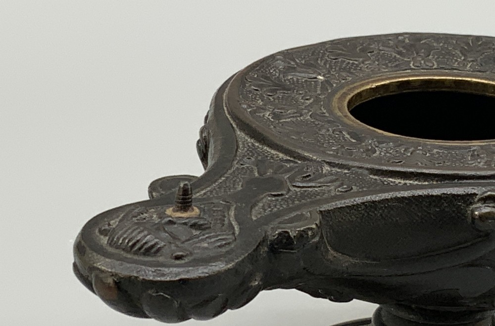 A patinated dark bronze classical oil lamp featuring a seated putto figure on top and a scroll handle resting on a pedestal base. View X