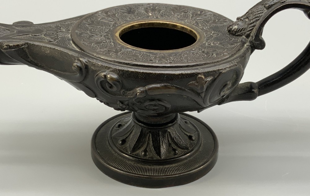 A patinated dark bronze classical oil lamp featuring a seated putto figure on top and a scroll handle resting on a pedestal base. View XI