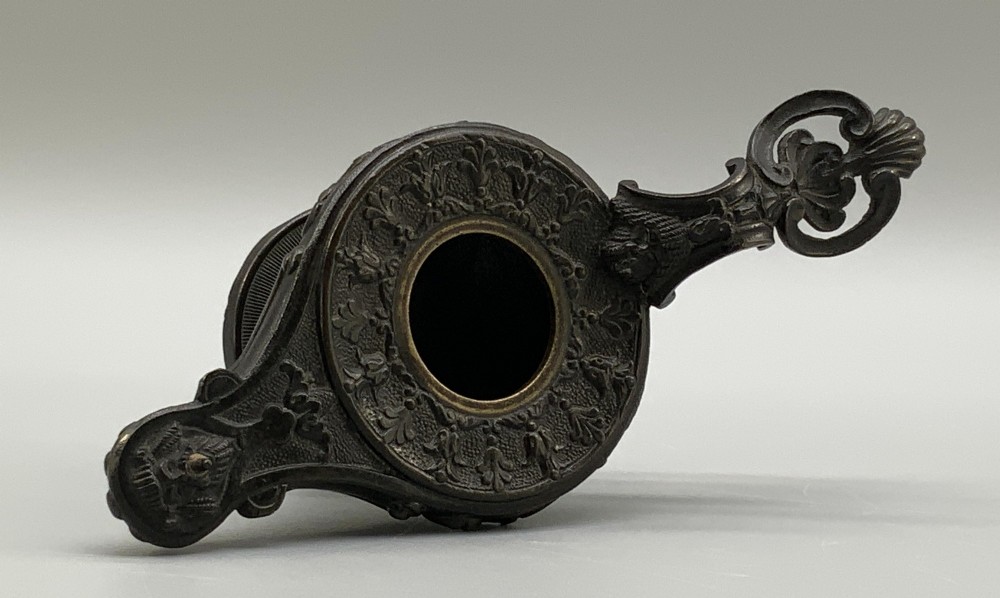A patinated dark bronze classical oil lamp featuring a seated putto figure on top and a scroll handle resting on a pedestal base. View XII