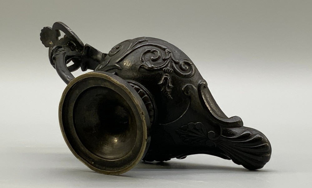 A patinated dark bronze classical oil lamp featuring a seated putto figure on top and a scroll handle resting on a pedestal base. View XIII