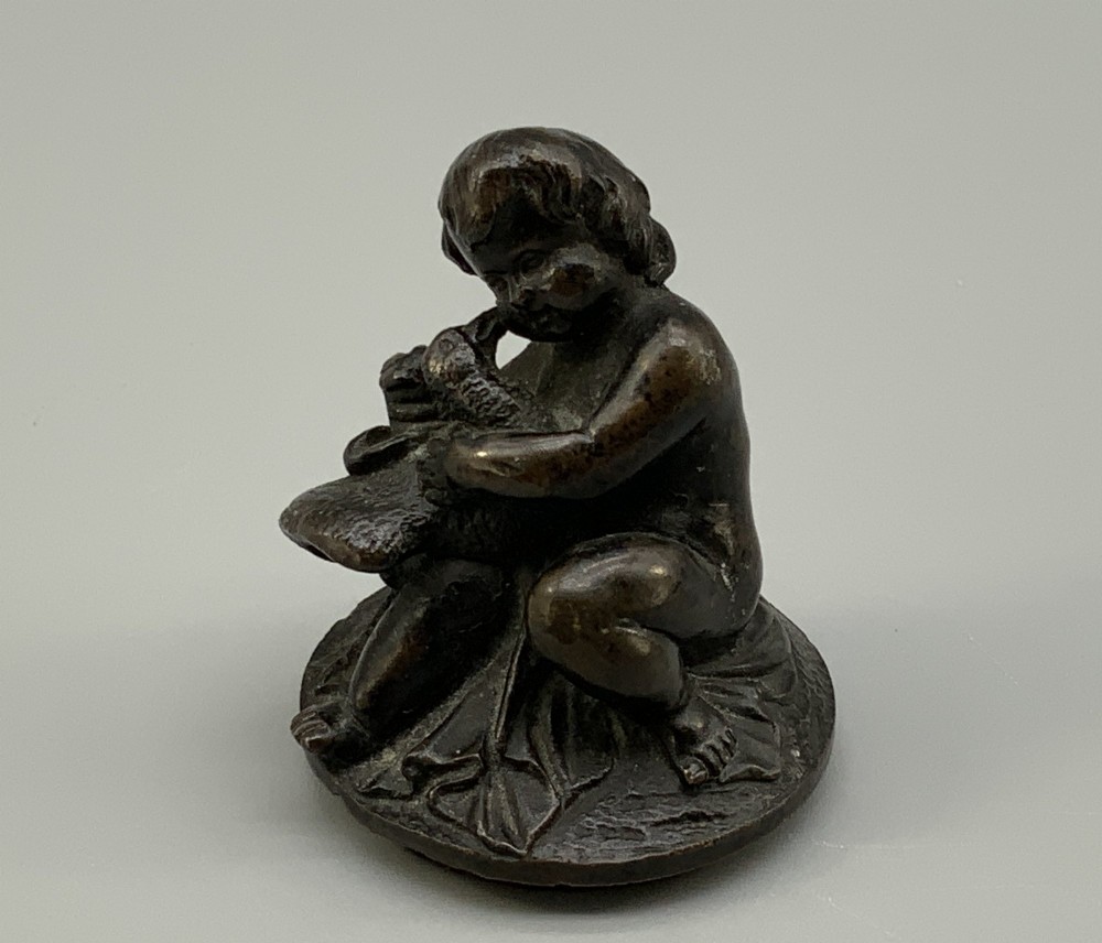 A patinated dark bronze classical oil lamp featuring a seated putto figure on top and a scroll handle resting on a pedestal base. View XIV