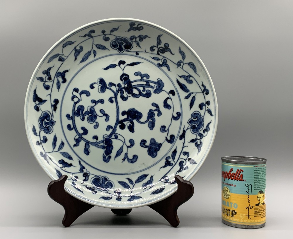 A circular Chinese blue and white porcelain charger decorated with hand-painted scrolling foliage and floral motifs, resting on a dark wooden stand. View III