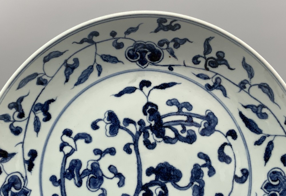 A circular Chinese blue and white porcelain charger decorated with hand-painted scrolling foliage and floral motifs, resting on a dark wooden stand. View IV