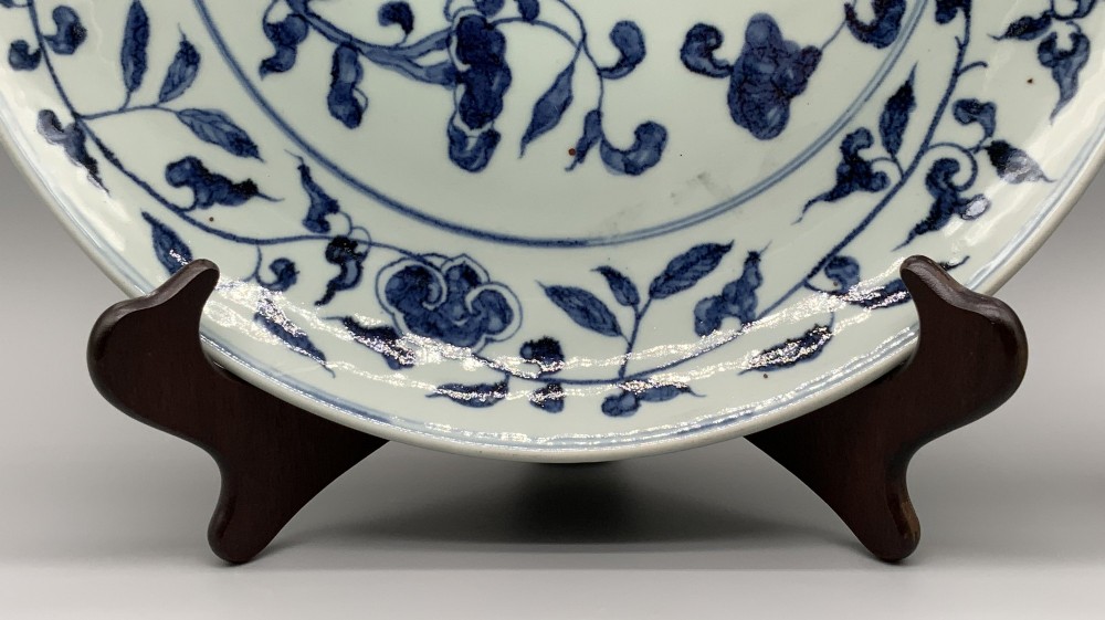 A circular Chinese blue and white porcelain charger decorated with hand-painted scrolling foliage and floral motifs, resting on a dark wooden stand. View VII