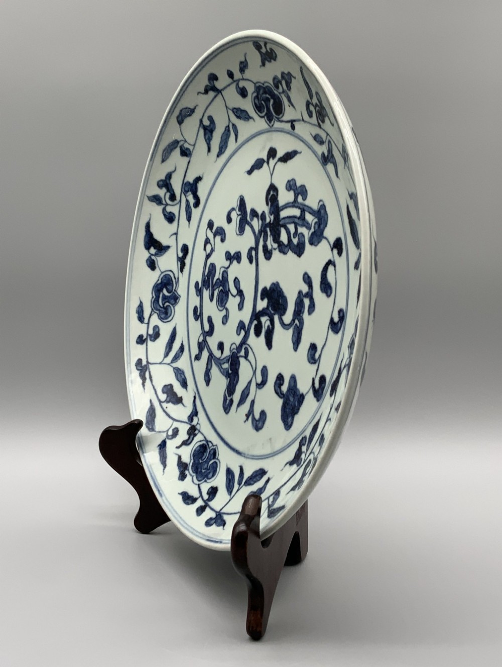 A circular Chinese blue and white porcelain charger decorated with hand-painted scrolling foliage and floral motifs, resting on a dark wooden stand. View X