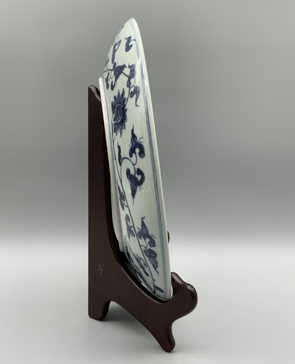A circular Chinese blue and white porcelain charger decorated with hand-painted scrolling foliage and floral motifs, resting on a dark wooden stand. View XI