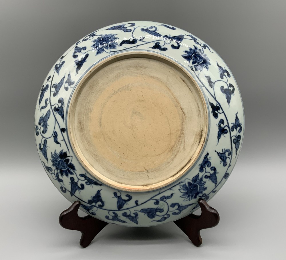A circular Chinese blue and white porcelain charger decorated with hand-painted scrolling foliage and floral motifs, resting on a dark wooden stand. View XII