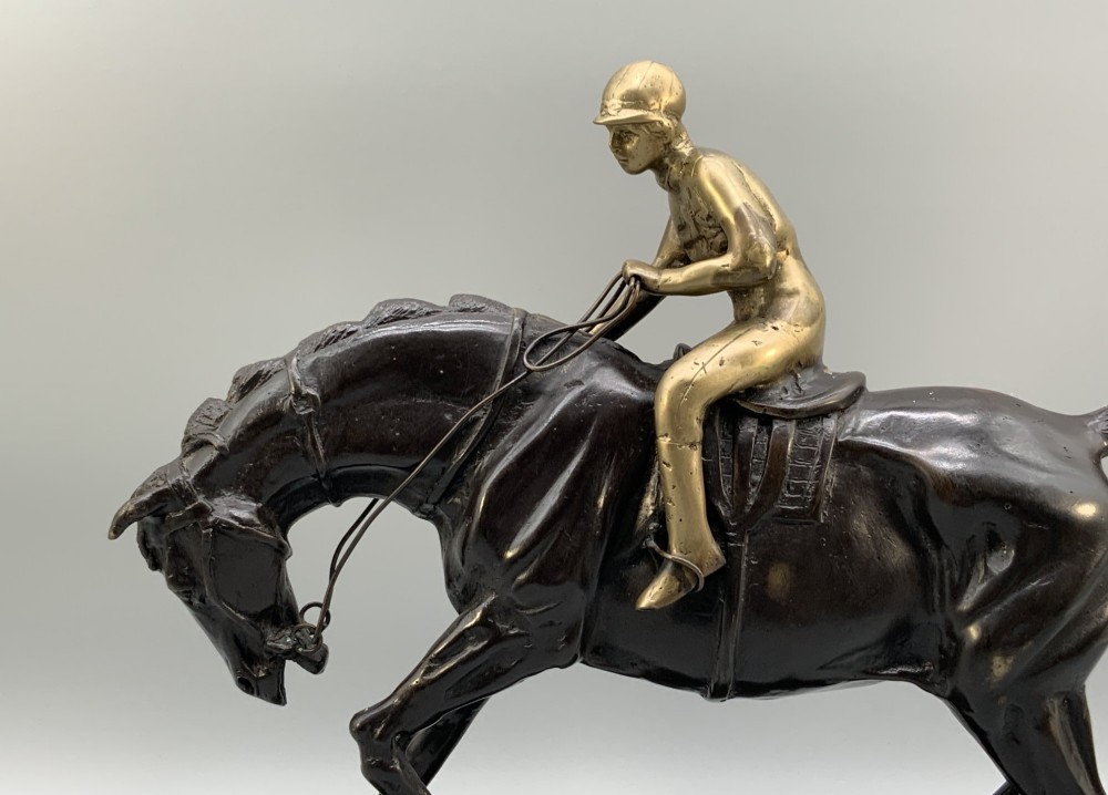 A patinated bronze sculpture depicting a racehorse and jockey mid-stride set upon a textured rectangular bronze base. View V