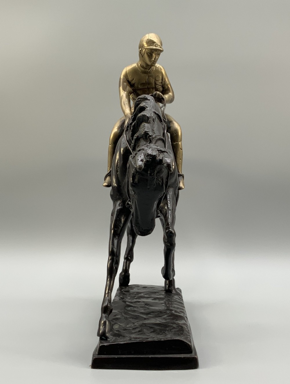 A patinated bronze sculpture depicting a racehorse and jockey mid-stride set upon a textured rectangular bronze base. View VI