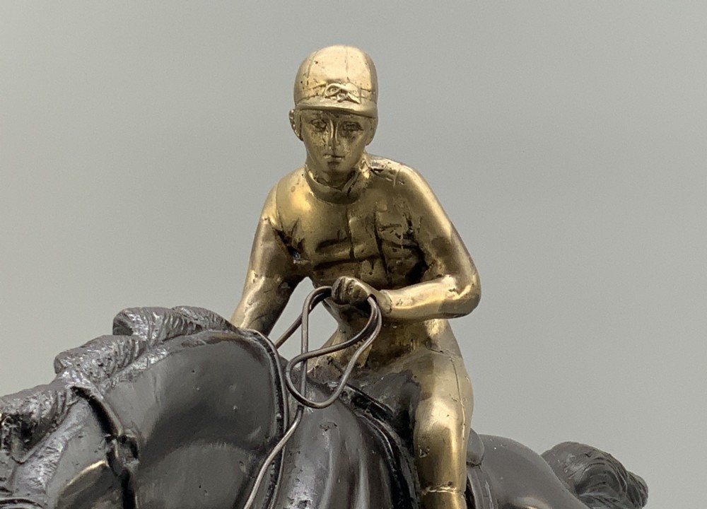 A patinated bronze sculpture depicting a racehorse and jockey mid-stride set upon a textured rectangular bronze base. View XIII