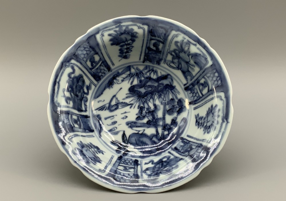 A circular porcelain bowl with a lobed edge, decorated in cobalt blue underglaze featuring a central landscape surrounded by panelled floral reserves.