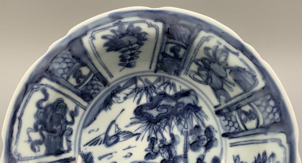 A circular porcelain bowl with a lobed edge, decorated in cobalt blue underglaze featuring a central landscape surrounded by panelled floral reserves. View V