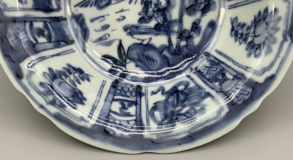 A circular porcelain bowl with a lobed edge, decorated in cobalt blue underglaze featuring a central landscape surrounded by panelled floral reserves. View VI