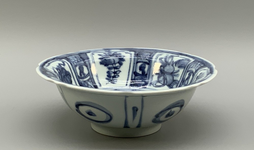 A circular porcelain bowl with a lobed edge, decorated in cobalt blue underglaze featuring a central landscape surrounded by panelled floral reserves. View IX