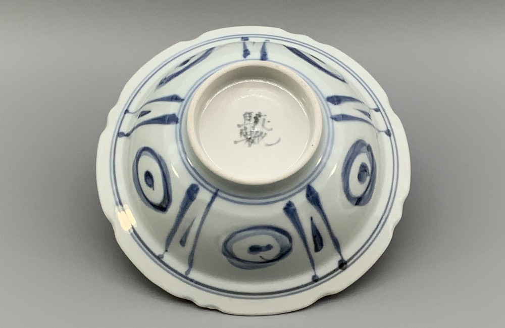 A circular porcelain bowl with a lobed edge, decorated in cobalt blue underglaze featuring a central landscape surrounded by panelled floral reserves. View X