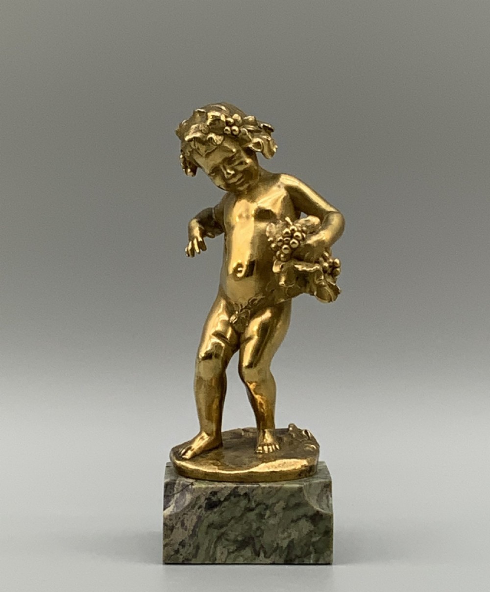A small gilt bronze sculpture depicting an infant Bacchus holding a bunch of grapes, set upon a square green marble plinth.