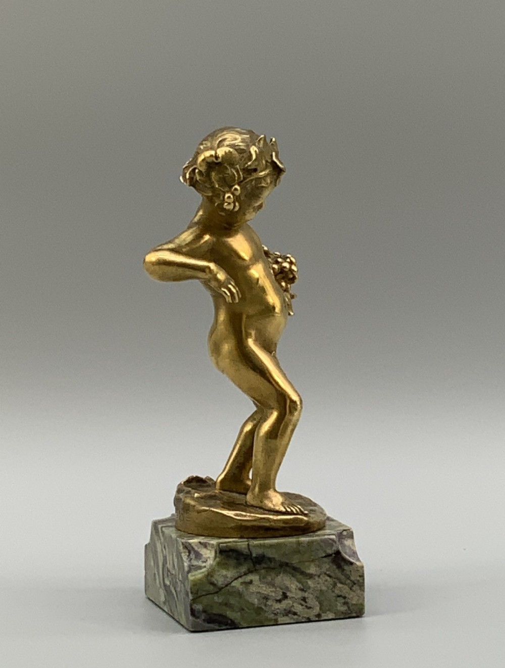 A small gilt bronze sculpture depicting an infant Bacchus holding a bunch of grapes, set upon a square green marble plinth. View VIII