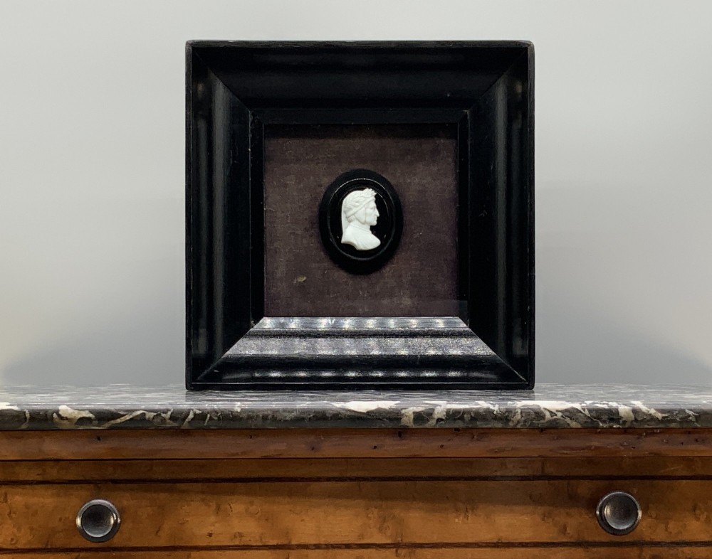 A small carved white classical profile portrait cameo mounted on a dark background within a deep black ebonised square frame.
