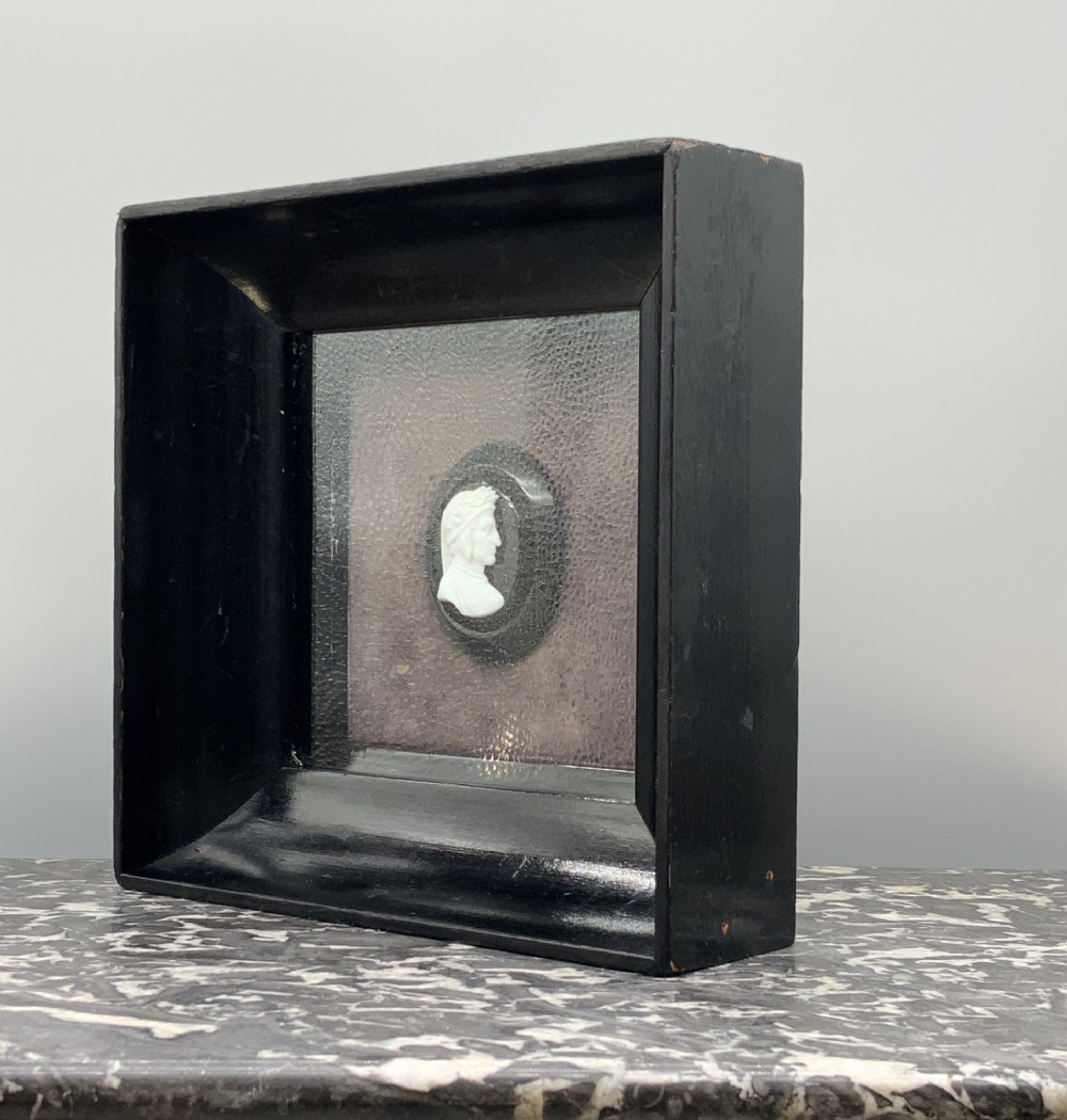 A small carved white classical profile portrait cameo mounted on a dark background within a deep black ebonised square frame. View VI