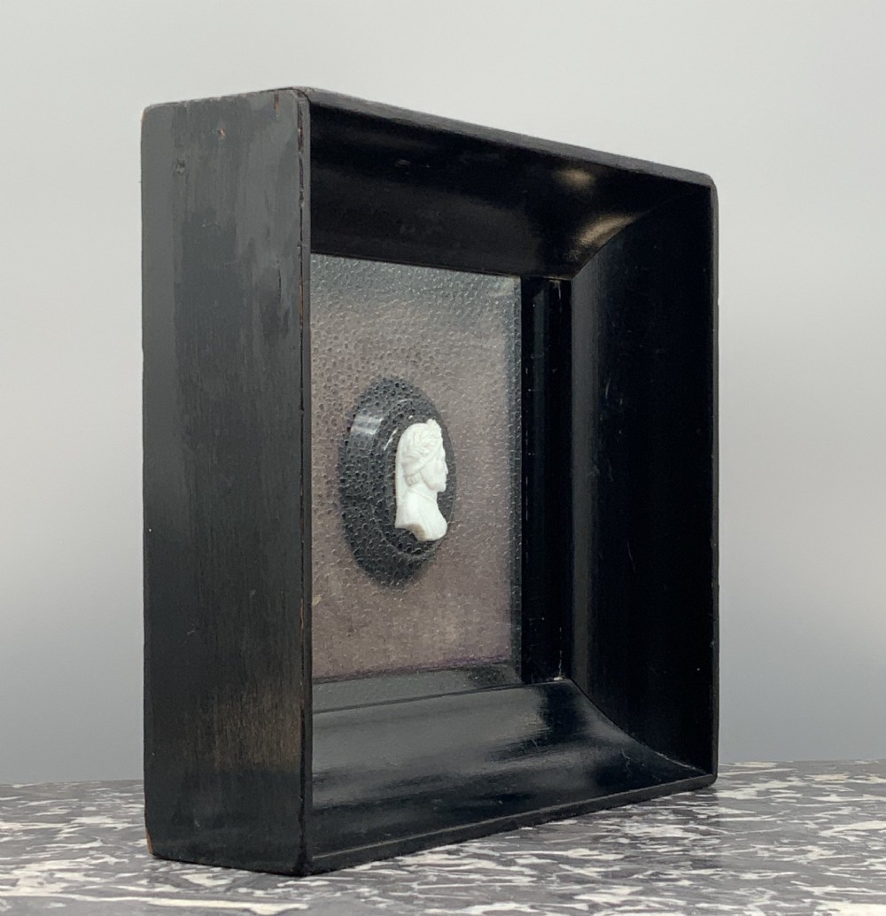 A small carved white classical profile portrait cameo mounted on a dark background within a deep black ebonised square frame. View VII