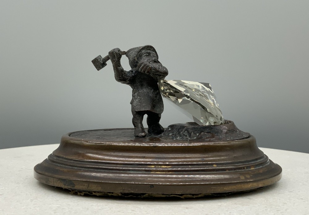 A small dark bronze figurine of a bearded dwarf miner holding a hammer overhead, standing on a molded base next to a faceted cut crystal gem.