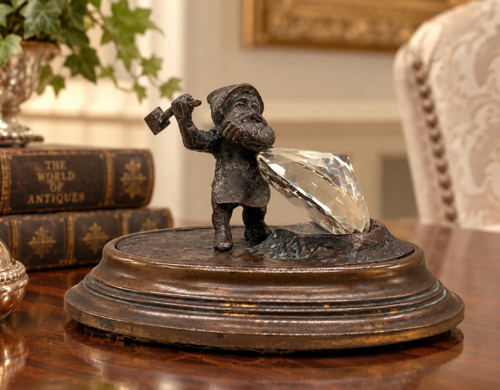 A small dark bronze figurine of a bearded dwarf miner holding a hammer overhead, standing on a molded base next to a faceted cut crystal gem. View II